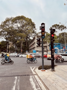 An urban street intersection with several motorbikes and cars waiting at a traffic light. Lush green trees tower on the roadside, and buildings line the street. A delivery rider on a motorbike carries a large box, while others are wearing helmets and navigating through the traffic. The sky is cloudy, providing a muted backdrop to the bustling scene.