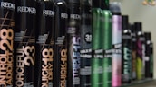 A row of various hair styling products in black cylindrical containers, with labels displaying product names such as 'Control Addict', 'Forceful', 'Quick Dry', and others. The bottles have a sleek design with some featuring metallic accents in colors like green and purple.