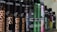 A row of various hair styling products in black cylindrical containers, with labels displaying product names such as 'Control Addict', 'Forceful', 'Quick Dry', and others. The bottles have a sleek design with some featuring metallic accents in colors like green and purple.
