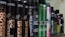 A row of various hair styling products in black cylindrical containers, with labels displaying product names such as 'Control Addict', 'Forceful', 'Quick Dry', and others. The bottles have a sleek design with some featuring metallic accents in colors like green and purple.