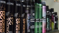 A row of various hair styling products in black cylindrical containers, with labels displaying product names such as 'Control Addict', 'Forceful', 'Quick Dry', and others. The bottles have a sleek design with some featuring metallic accents in colors like green and purple.