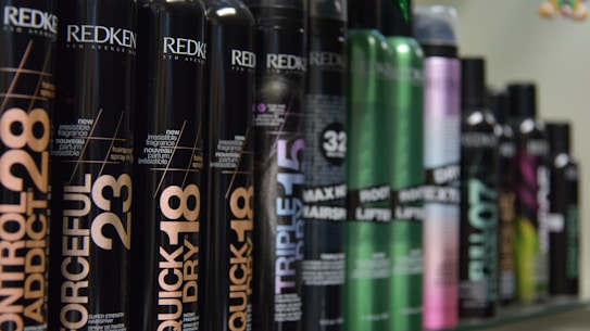 A row of various hair styling products in black cylindrical containers, with labels displaying product names such as 'Control Addict', 'Forceful', 'Quick Dry', and others. The bottles have a sleek design with some featuring metallic accents in colors like green and purple.