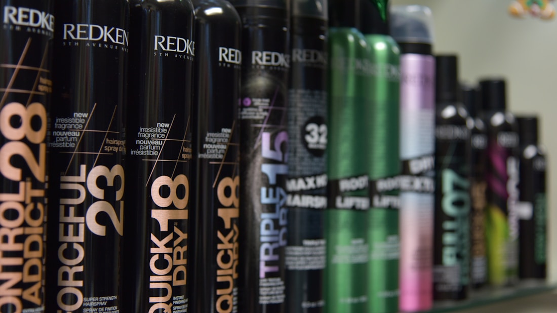 A row of various hair styling products in black cylindrical containers, with labels displaying product names such as 'Control Addict', 'Forceful', 'Quick Dry', and others. The bottles have a sleek design with some featuring metallic accents in colors like green and purple.
