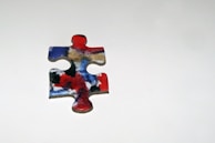 A single jigsaw puzzle piece with a colorful abstract design featuring red, blue, black, and white swirls, placed on a plain light gray background.