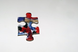A single jigsaw puzzle piece with a colorful abstract design featuring red, blue, black, and white swirls, placed on a plain light gray background.