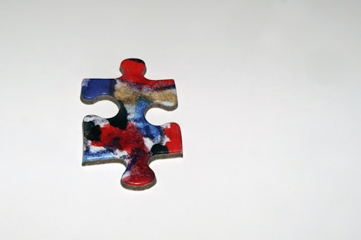 A single jigsaw puzzle piece with a colorful abstract design featuring red, blue, black, and white swirls, placed on a plain light gray background.