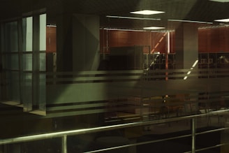 Interior space with large glass windows featuring horizontal translucent stripes. Inside, there are red blinds, and an office or study area is visible with tables and chairs. Metal railings are present in the foreground.