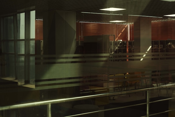 Interior space with large glass windows featuring horizontal translucent stripes. Inside, there are red blinds, and an office or study area is visible with tables and chairs. Metal railings are present in the foreground.