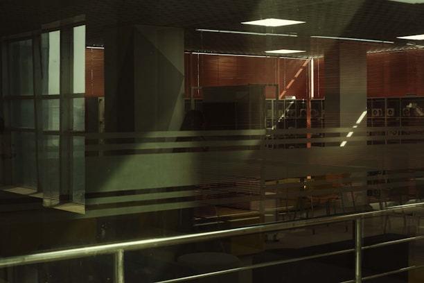 Interior space with large glass windows featuring horizontal translucent stripes. Inside, there are red blinds, and an office or study area is visible with tables and chairs. Metal railings are present in the foreground.