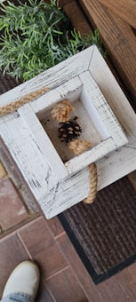 A close-up view of a rustic wooden decorative piece with distressed white paint. A pinecone and a thick rope are placed within the frame. In the background, there are green plants, brick tiles, and a doormat. A part of a shoe is visible at the bottom.