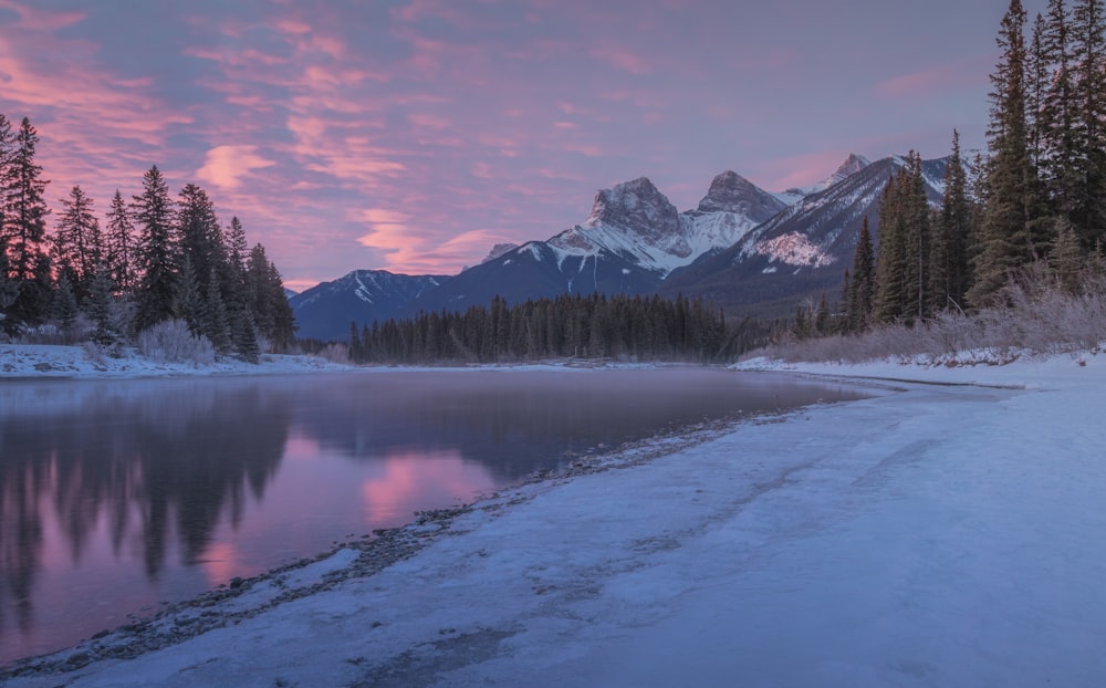 a lake surrounded by snow covered mountains under a pink sky