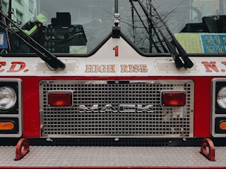The image features the front of a fire truck with the brand 'MACK' prominently displayed on the grill. Above the grill, the words 'HIGH RISE' are printed. The truck is painted in red and white, typical of emergency vehicles. The reflection of nearby buildings can be seen in the windshield, along with some books or manuals placed on the dashboard. Part of a cityscape with leafless tree branches is visible through the transparent parts.