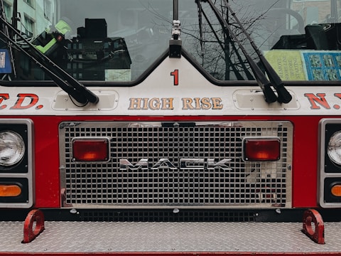 The image features the front of a fire truck with the brand 'MACK' prominently displayed on the grill. Above the grill, the words 'HIGH RISE' are printed. The truck is painted in red and white, typical of emergency vehicles. The reflection of nearby buildings can be seen in the windshield, along with some books or manuals placed on the dashboard. Part of a cityscape with leafless tree branches is visible through the transparent parts.