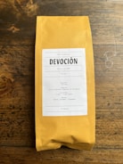 A package of coffee labeled 'DEVOCIÓN', featuring details about its Colombian origin, including variety, region, altitude, and flavor notes. The package is placed on a rustic wooden surface.