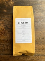 A package of coffee labeled 'DEVOCIÓN', featuring details about its Colombian origin, including variety, region, altitude, and flavor notes. The package is placed on a rustic wooden surface.