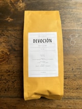 A package of coffee labeled 'DEVOCIÓN', featuring details about its Colombian origin, including variety, region, altitude, and flavor notes. The package is placed on a rustic wooden surface.