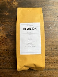 A package of coffee labeled 'DEVOCIÓN', featuring details about its Colombian origin, including variety, region, altitude, and flavor notes. The package is placed on a rustic wooden surface.