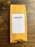 A package of coffee labeled 'DEVOCI&Oacute;N', featuring details about its Colombian origin, including variety, region, altitude, and flavor notes. The package is placed on a rustic wooden surface.