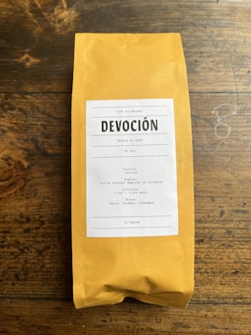 A package of coffee labeled 'DEVOCIÓN', featuring details about its Colombian origin, including variety, region, altitude, and flavor notes. The package is placed on a rustic wooden surface.