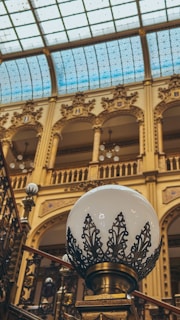 An intricately designed indoor setting featuring a large, ornate lamp with a decorative metal pattern surrounding the globe. The architecture includes arched windows and detailed moldings on the walls, with a glass ceiling that allows natural light to illuminate the space.