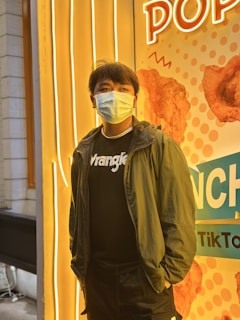 A person wearing a face mask and casual clothing stands in front of a brightly lit advertisement featuring images of fried chicken. The background includes neon lights and colorful graphics.