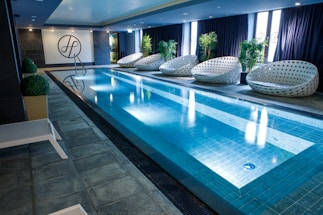 A sleek indoor swimming pool with clear blue water is surrounded by several modern, white woven lounge chairs arranged alongside the pool. Green potted plants add a touch of nature, while a stylish dark tile floor complements the contemporary design. The pool area is well-lit with overhead lights, and the pool ladder invites guests for a swim.