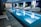 A sleek indoor swimming pool with clear blue water is surrounded by several modern, white woven lounge chairs arranged alongside the pool. Green potted plants add a touch of nature, while a stylish dark tile floor complements the contemporary design. The pool area is well-lit with overhead lights, and the pool ladder invites guests for a swim.
