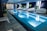 A sleek indoor swimming pool with clear blue water is surrounded by several modern, white woven lounge chairs arranged alongside the pool. Green potted plants add a touch of nature, while a stylish dark tile floor complements the contemporary design. The pool area is well-lit with overhead lights, and the pool ladder invites guests for a swim.