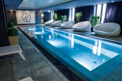 A sleek indoor swimming pool with clear blue water is surrounded by several modern, white woven lounge chairs arranged alongside the pool. Green potted plants add a touch of nature, while a stylish dark tile floor complements the contemporary design. The pool area is well-lit with overhead lights, and the pool ladder invites guests for a swim.