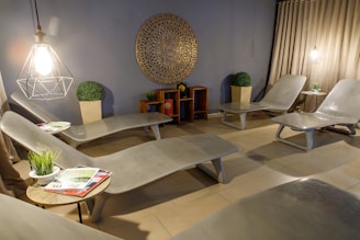 A serene and modern lounge area with several reclining chairs arranged around the room. The walls are adorned with a large circular decorative piece, and potted plants add a touch of greenery. Soft lighting from hanging lamps creates a warm ambiance. Small side tables hold plants and magazines, enhancing the inviting atmosphere.