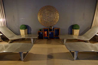 Four beige lounge chairs are symmetrically arranged in a room with a modern aesthetic. A decorative circular wall piece hangs centrally on a gray wall above two small shelves, each containing a potted plant and decorative items. Two large framed artificial plants sit on either side.