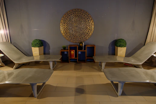 Four beige lounge chairs are symmetrically arranged in a room with a modern aesthetic. A decorative circular wall piece hangs centrally on a gray wall above two small shelves, each containing a potted plant and decorative items. Two large framed artificial plants sit on either side.