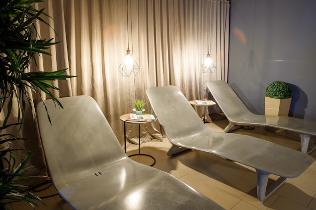 The image shows a relaxing lounge area with three modern, curved, gray lounge chairs. Each chair is accompanied by a small round table, one with a potted plant and reading material on it. The area is softly lit by two pendant lights with geometric wire frames, casting a warm glow on the beige curtain behind the chairs. There is a green artificial plant in a square pot in the background and some greenery on the left side of the image.