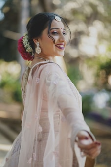 A woman is wearing a traditional outfit with intricate embroidery, adorned with jewelry including earrings, a necklace, and a decorative piece on her head. Her hair is styled elegantly with flowers. She has a joyful expression and her hand is extended towards the camera, suggesting a playful or inviting gesture. The background is softly blurred, enhancing the focus on her expression and attire.