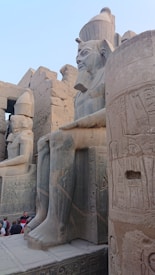Large stone statues of pharaohs are adorned with detailed inscriptions and carvings. The ancient architecture features hieroglyphics on the walls, and a group of tourists can be seen admiring the structure.
