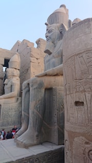 Large stone statues of pharaohs are adorned with detailed inscriptions and carvings. The ancient architecture features hieroglyphics on the walls, and a group of tourists can be seen admiring the structure.