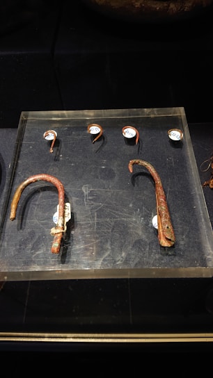 A collection of ancient, rusted metal artifacts are displayed on a dark surface. Two large, curved metal pieces rest on a clear platform at the bottom, while four smaller, semi-circular metal pieces with labels are positioned above them.