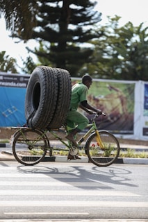 A man riding a bicycle is carrying two large tires secured vertically on the back of the bike. The man is wearing a green uniform, suggesting he might be involved in manual labor or transport work. The background includes large trees and a billboard with blurred imagery.