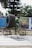 A man riding a bicycle is carrying two large tires secured vertically on the back of the bike. The man is wearing a green uniform, suggesting he might be involved in manual labor or transport work. The background includes large trees and a billboard with blurred imagery.