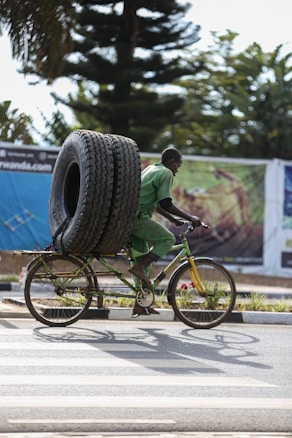 A man riding a bicycle is carrying two large tires secured vertically on the back of the bike. The man is wearing a green uniform, suggesting he might be involved in manual labor or transport work. The background includes large trees and a billboard with blurred imagery.
