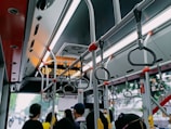 Inside view of a public bus featuring overhead handrails and handles for standing passengers. Several people are partially visible, facing towards the front of the bus. A digital sign indicates the next stop and general bus maintenance equipment can be observed.