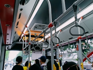 Inside view of a public bus featuring overhead handrails and handles for standing passengers. Several people are partially visible, facing towards the front of the bus. A digital sign indicates the next stop and general bus maintenance equipment can be observed.