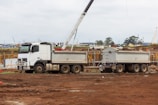 A white construction truck with two trailers is parked on a dirt construction site. In the background, a crane is visible, and there are construction materials and machinery scattered around. The sky is overcast, and the site has a busy, industrial atmosphere.