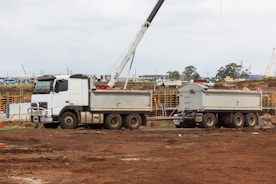 A white construction truck with two trailers is parked on a dirt construction site. In the background, a crane is visible, and there are construction materials and machinery scattered around. The sky is overcast, and the site has a busy, industrial atmosphere.