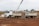 A white construction truck with two trailers is parked on a dirt construction site. In the background, a crane is visible, and there are construction materials and machinery scattered around. The sky is overcast, and the site has a busy, industrial atmosphere.