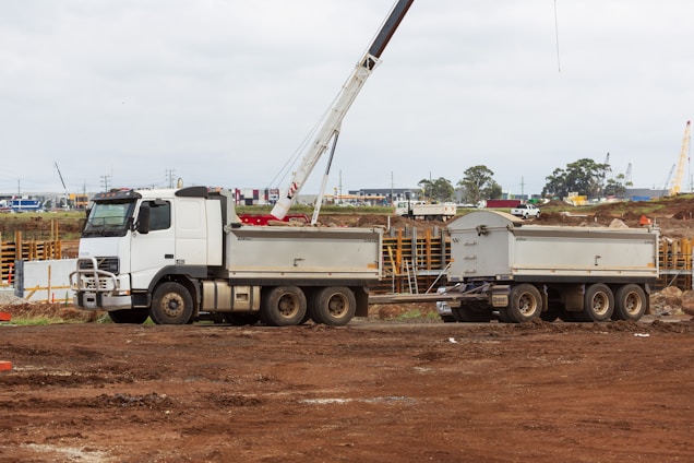 A white construction truck with two trailers is parked on a dirt construction site. In the background, a crane is visible, and there are construction materials and machinery scattered around. The sky is overcast, and the site has a busy, industrial atmosphere.