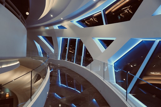A futuristic interior features a curved walkway with sleek, white walls punctuated by geometric, angular windows. The windows are lined with blue LED lights, creating a modern and dynamic atmosphere. The reflective floor mirrors the bright lighting, enhancing the overall sleek aesthetic.