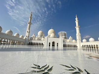 A grand mosque with stunning white domes and intricate minarets stretches across the horizon, gleaming under a bright blue sky. Ornamental details and symmetrical arches contribute to the architectural elegance. The expansive marble courtyard reflects the sunlight, adding to the serene and majestic atmosphere.