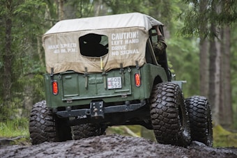 A rugged off-road vehicle with large tires is navigating through a muddy forest trail. The vehicle has a canvas cover with printed warnings about speed and signaling, and a visible license plate. Surrounding the vehicle, a dense forest with tall trees and lush greenery adds to the adventurous atmosphere.