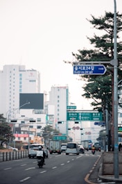 An urban street scene with a variety of vehicles including cars, a motorcycle, and buses, moving along a multi-lane road. Tall office buildings and street signs with Korean and English text are visible in the background, with green and red traffic lights overhead. Trees line the street, adding greenery to the city landscape.